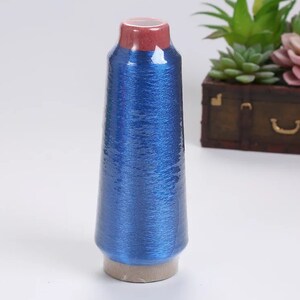 May include: A spool of blue metallic thread, wrapped in clear plastic. The top of the spool is a deep red color. The thread is a vibrant blue, reflecting light. The spool sits on a light brown base. Decorative plants and a small wooden chest are in the background.