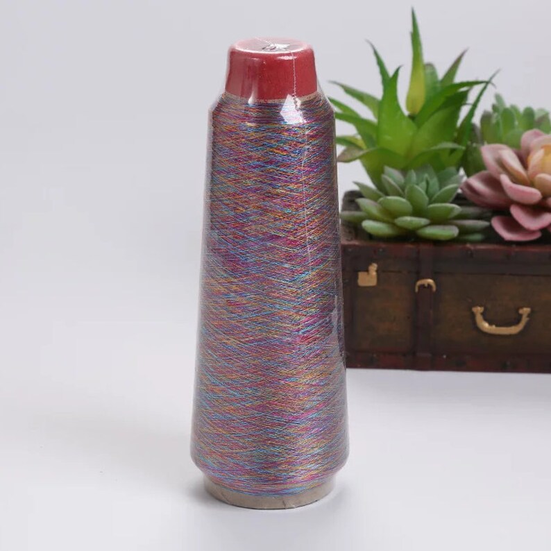 May include: A cone of colorful yarn wrapped in clear plastic. The yarn has a rainbow of colors including blue, purple, yellow, and red. The top of the cone is a deep red color. The yarn is on a white surface.