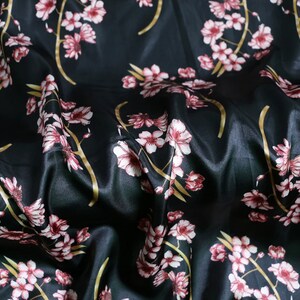 K&ouml;nnte beinhalten: Schwarzer Satin-Stoff mit floralem Muster. Das Design zeigt rosa und wei&szlig;e Bl&uuml;ten mit gelben Stielen. Der Stoff ist drapiert und erzeugt weiche Falten und Schatten. Der Gesamteffekt ist elegant und luxuri&ouml;s.