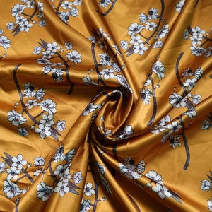 K&ouml;nnte beinhalten: Senffarbener Satin-Stoff mit floralem Muster. Das Design zeigt wei&szlig;e Bl&uuml;ten und dunkelgraue Zweige. Der Stoff ist drapiert, wodurch Falten und Schatten entstehen, die seinen Glanz hervorheben. Ideal f&uuml;r Kleidung oder Heimdekoration.