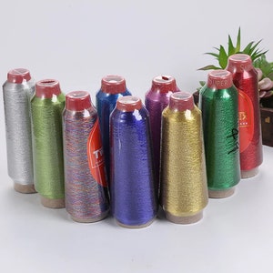 May include: A collection of colorful metallic thread spools. The spools are wrapped in clear plastic and feature a variety of colors, including silver, green, blue, purple, and gold. Each spool has a red cap. The text "SUPER" is visible on some labels.