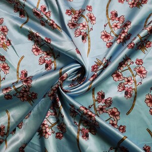 K&ouml;nnte beinhalten: Hellblauer Satin-Stoff mit floralem Muster. Das Design zeigt rosa Bl&uuml;ten und braune Stiele. Der Stoff ist drapiert und zeigt den Glanz und die Textur des Materials.