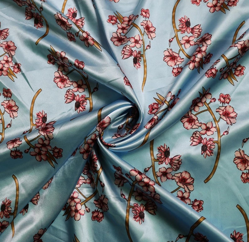 K&ouml;nnte beinhalten: Hellblauer Satin-Stoff mit floralem Muster. Das Design zeigt rosa Bl&uuml;ten und braune Stiele. Der Stoff ist drapiert und zeigt den Glanz und die Textur des Materials.