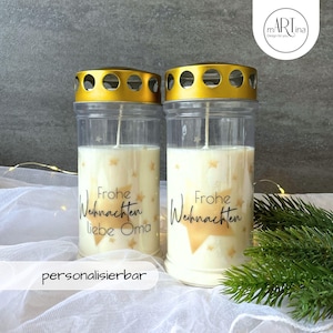 Könnte beinhalten: Zwei gläserne Kerzenhalter mit goldenen Deckeln und weißen Kerzen. Die Kerzen haben ein goldenes Sternendesign und den Text "Frohe Weihnachten" und "liebe Oma".