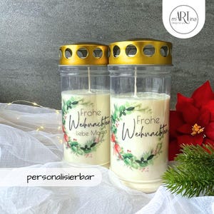 Könnte beinhalten: Zwei klare Glaskerzenbehälter mit goldenen Deckeln und weißen Kerzen. Die Behälter haben ein grünes und weißes Blumenmuster mit dem Text "Frohe Weihnachten" und "liebe Mama".