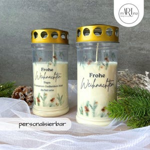 Könnte beinhalten: Zwei klare Glas-Kerzenhalter mit goldenen Deckeln und weißen Kerzen. Die Kerzen haben ein gedrucktes Design aus Kiefernästen und den Text "Frohe Weihnachten". Die Kerze links hat außerdem den Text "Papa" und "In unseren Gedanken bist du bei uns".