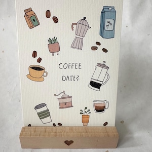 Könnte beinhalten: Eine rechteckige Grußkarte mit dem Text COFFEE DATE?, umgeben von Illustrationen von Kaffeebohnen, einem Espressokocher, einer French Press, einer Kaffeemühle, Tassen und Topfpflanzen, präsentiert auf einem hellen Holzständer mit Herzausschnitt.