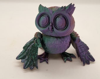 Puede incluir: Una figurita de búho iridiscente de color púrpura y verde con alas detalladas y ojos grandes. El búho está hecho de un material texturizado y tiene un estilo caprichoso y de dibujos animados.