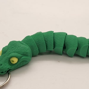 以下が含まれることがあります： 黄色の目をした緑色のセグメント化されたヘビのキーホルダー。ヘビは丸まっており、頭にリングが付いています。