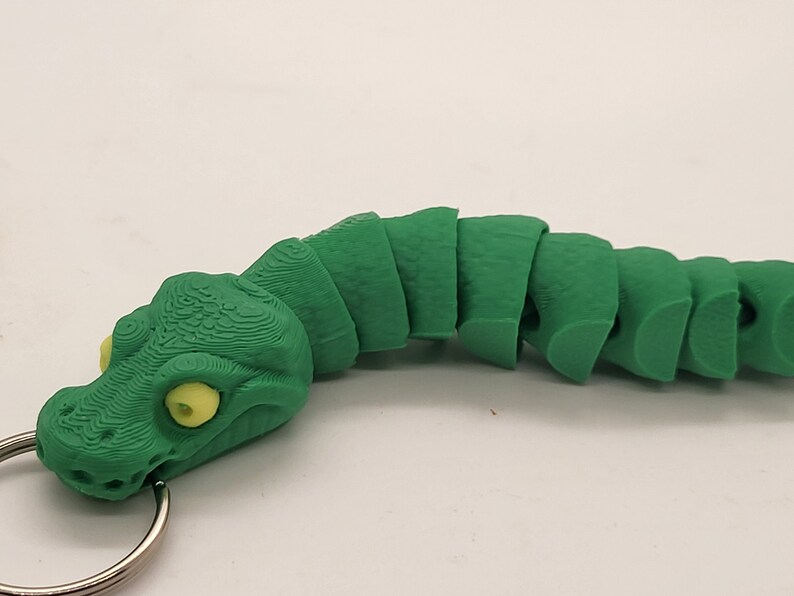 以下が含まれることがあります： 黄色の目をした緑色のセグメント化されたヘビのキーホルダー。ヘビは丸まっており、頭にリングが付いています。