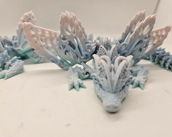 May include: Three intricately detailed, 3D printed dragon figurines with delicate, lacy wings. The dragons are painted in a gradient of light blue and white, with hints of pink.