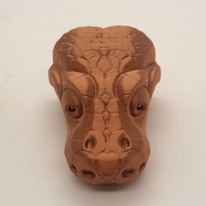 Può includere: Una scultura di testa di alligatore in 3D, di colore marrone, con occhi dettagliati e pelle testurizzata.