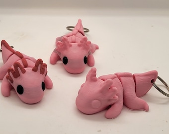 May include: Three pink axolotl keychains with silver key rings. The axolotls are in different poses, with one lying down, one standing up, and one swimming.