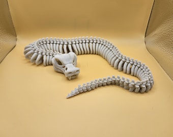May include: A light gray, articulated snake skeleton model. The model features a detailed skull and spine, with each vertebra visible. The snake is posed in a curved position, suggesting movement. The model is made of a light gray material.