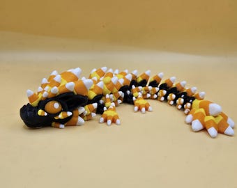 May include: A 3D-printed dragon figurine in black, orange, yellow, and white. The dragon's body is segmented, with candy corn-shaped spikes along its back. The head has large eyes and detailed features. The figurine is on a solid yellow background.