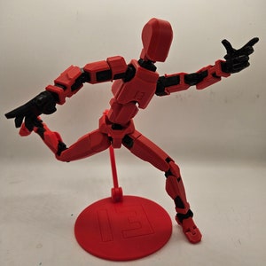 May include: A red articulated action figure with a black base. The figure is posed in a dynamic stance with one leg raised and the other leg bent. The figure has a simple design with no facial features.