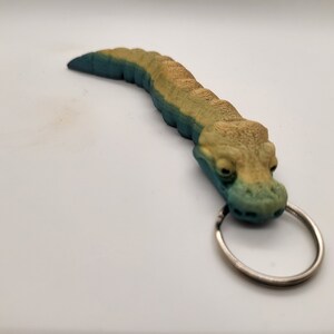 以下が含まれることがあります： 銀色のリングが付いた緑と黄色のワニのキーホルダー。ワニは質感のある鱗で詳細に作られており、リアルな外観をしています。