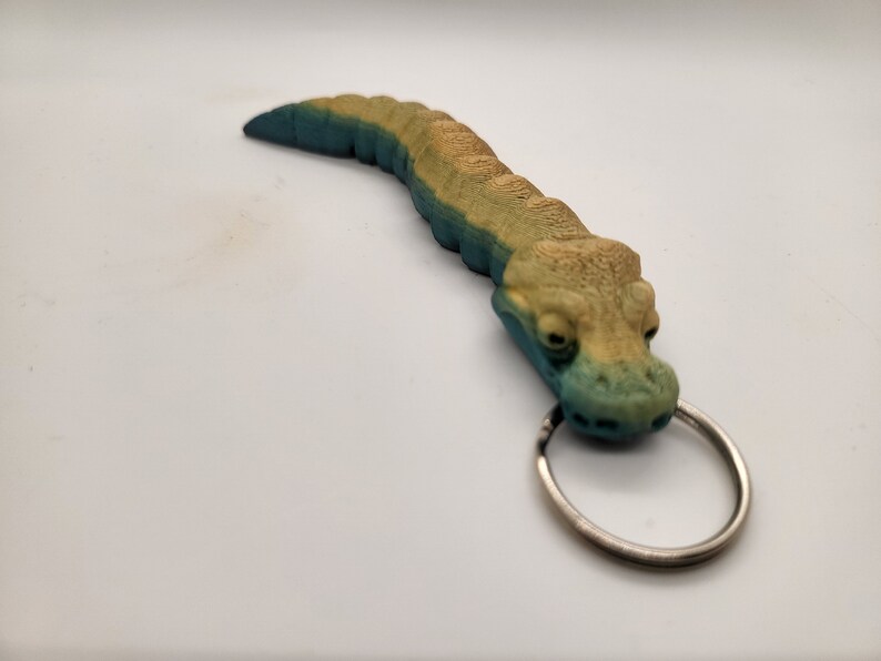 以下が含まれることがあります： 銀色のリングが付いた緑と黄色のワニのキーホルダー。ワニは質感のある鱗で詳細に作られており、リアルな外観をしています。