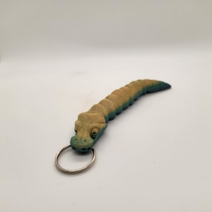 Może przedstawiać: Brelok do kluczy w kształcie zielonego i brązowego aligatora z srebrnym kółkiem. Aligator jest szczegółowo wykonany z fakturowanych łusek i ma realistyczny wygląd.