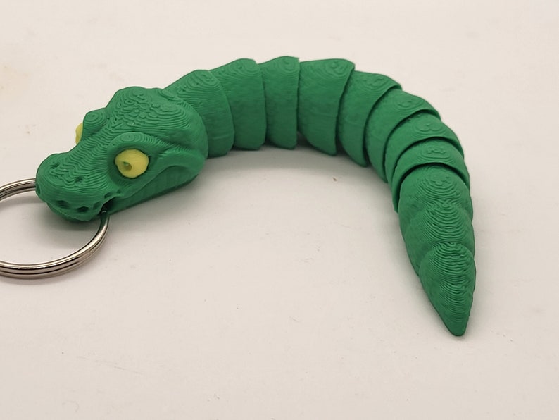 以下が含まれることがあります： キーリングが付いた緑色のプラスチック製のヘビのキーホルダー。ヘビはセグメント化されたボディと黄色の目をしています。