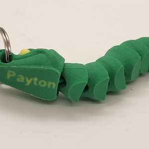 以下が含まれることがあります： 銀色のキーリングと頭部に「Payton」という名前が印刷された、緑色のセグメント化された毛虫のキーホルダーです。