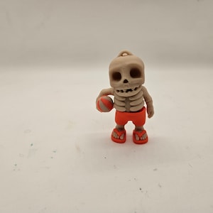 Peut inclure: Une petite figurine de squelette beige portant un short de bain orange et tenant un petit ballon orange. La figurine est debout sur une surface blanche.