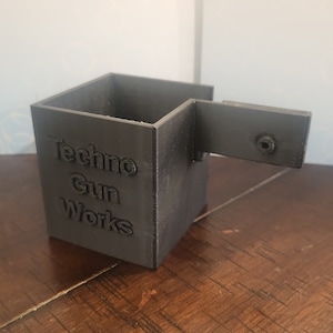 Può includere: Una scatola grigia stampata in 3D con un coperchio incernierato. La scatola ha le parole "Techno Gun Works" stampate sul davanti.