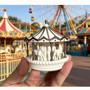 May include: A miniature carousel figurine with white horses and a black and white striped canopy. The base and horses are white, and the support poles are black. The background shows a blurred amusement park scene with a Ferris wheel.