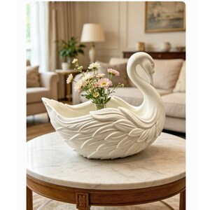May include: A white ceramic swan-shaped planter filled with pink and yellow flowers. The planter sits on a round marble-topped table with a wooden base. The swan has detailed feather and wing designs.