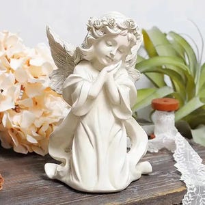 Puede incluir: Una figura de ángel color crema con las manos juntas y una corona floral. El ángel tiene alas detalladas y está arrodillado. El fondo incluye flores y vegetación, creando una escena serena y decorativa.