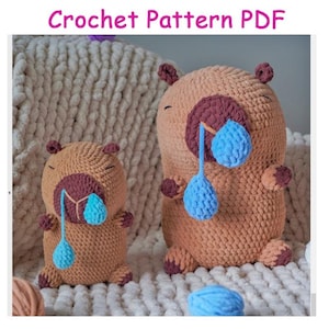 May include: Two crocheted bear-like figures, one large and one small, with brown bodies, dark burgundy noses, and blue teardrops. The text "Crochet Pattern PDF" is at the top. The figures are on a cream-colored, textured surface.