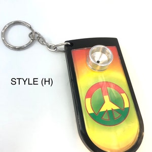 May include: A black keyring featuring a rectangular body with rounded edges and a metal ring. The centre displays a gradient background of red, yellow, and green with a peace symbol. A silver metal bowl is attached to the top. Text reads STYLE (H).