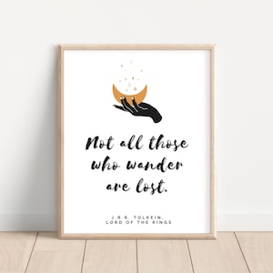 Può includere: Una stampa incorniciata bianca con una mano nera che tiene una luna crescente dorata con stelle. La stampa presenta la citazione "Non tutti coloro che vagano sono perduti." di J.R.R. Tolkien, autore de Il Signore degli Anelli.