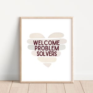 May include: A framed print with the words "WELCOME PROBLEM SOLVERS" in burgundy text, arranged in a heart shape. The heart is composed of layered, abstract shapes in neutral tones. The frame is light wood.
