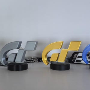 Puede incluir: Cuatro trofeos con el logo GT en colores sobre bases negras. Los trofeos son de bronce, plata, oro y una combinación azul y roja. El logo es una letra G y T estilizada. El logo de F44 Designs es visible en el fondo.