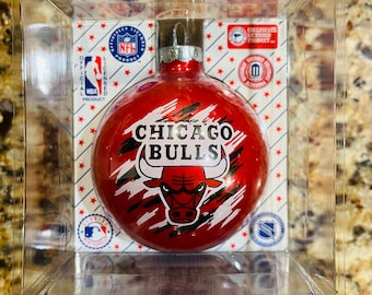 Pode incluir: Uma bola de Natal vermelha dos Chicago Bulls em uma caixa de plástico transparente. O enfeite apresenta o logotipo e o texto da equipe, com logotipos oficiais de produtos licenciados da NFL, NBA, MLB e NHL. O fundo tem um padrão de estrelas vermelhas.