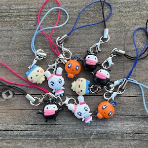 Puede incluir: Una colección de coloridos colgantes de personajes con cordones adjuntos. Los colgantes presentan varios diseños, incluyendo un conejo, una vaca y otras figuras de dibujos animados. Los cordones son de color azul, rosa y negro, con cierres plateados. Los colgantes están sobre una superficie de madera.