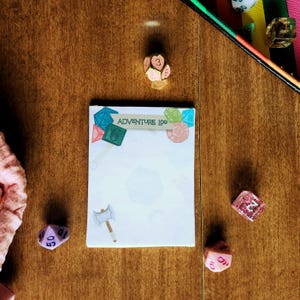 May include: A rectangular notepad with the text ADVENTURE LOG printed at the top, decorated with illustrations of multi-sided dice and a battle axe. The notepad is surrounded by several polyhedral dice in various colors and finishes.