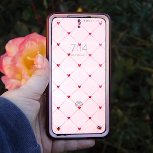 Könnte beinhalten: Ein Smartphone mit rosa Hülle zeigt einen rosa Hintergrund mit einem Muster aus roten Herzen. Die Uhrzeit 7:14 und das Datum Samstag, 7. Februar sind sichtbar. Das Telefon wird in einer Hand gehalten.