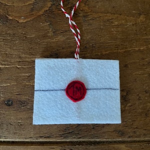May include: A white felt envelope with a red wax seal and a red and white string. The seal has the initials "IM" in the centre. The envelope has a black line drawn across the middle.