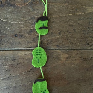 May include: A handmade green felt garland featuring three bean-shaped faces with brown hair and moustaches. The faces are connected by a green string, and the top has a loop for hanging. The garland is a whimsical decoration.