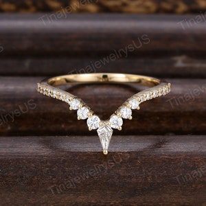 May include: A gold-colored ring with a V-shaped design. The ring features small, clear gemstones along the band and larger, clear gemstones at the center. The ring is set against a dark brown wooden surface.