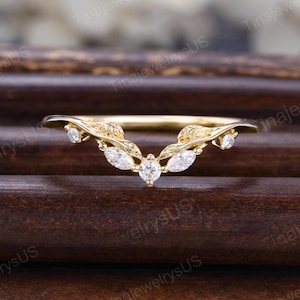 May include: A gold V-shaped ring featuring a delicate leaf-inspired design along the band. The ring is accented with five clear, sparkling stones, including two marquise-cut stones, one round-cut center stone, and two smaller round-cut side stones.