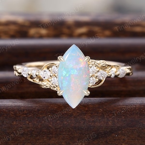 May include: A gold ring featuring a marquise-cut opal gemstone with iridescent colors. The ring has a delicate design with small, clear gemstones and leaf-like details on the band. The ring is set against a dark wooden background.