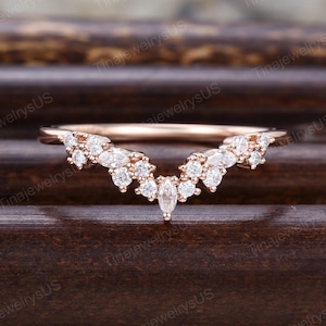 May include: Rose gold wedding band with a curved design. The band is adorned with multiple round and marquise-cut clear gemstones, creating a sparkling, elegant look. The ring is set against a dark wooden surface.
