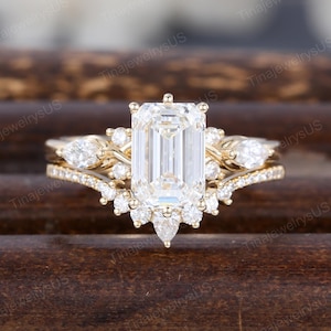 May include: A gold engagement ring set featuring an emerald-cut center stone and a delicate band with smaller accent stones. The ring is set against a dark wooden background, showcasing its intricate design and sparkle.