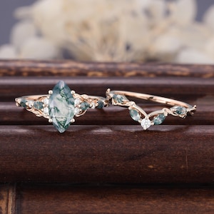 May include: A rose gold ring set featuring a marquise-cut moss agate center stone flanked by smaller round diamonds and marquise-cut moss agate side stones, paired with a matching V-shaped band adorned with marquise-cut moss agate and diamonds.