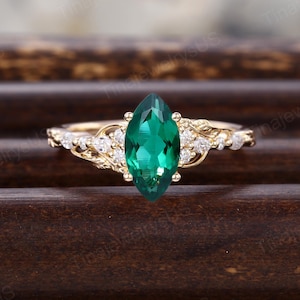 May include: A gold ring with a marquise-cut emerald center stone. The ring features a delicate design with small, clear accent stones and intricate detailing along the band. The emerald is a vibrant green color, and the ring is set against a dark wooden surface.