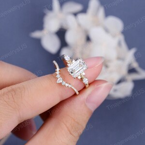 May include: A gold engagement ring set featuring a large emerald-cut diamond center stone. The ring is accompanied by a curved band with smaller accent stones. The rings are displayed against a soft, blurred background.