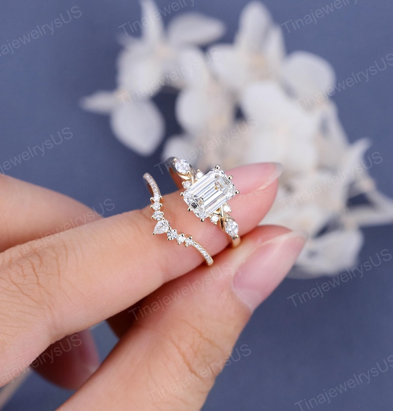 May include: A gold engagement ring set featuring a large emerald-cut diamond center stone. The ring is accompanied by a curved band with smaller accent stones. The rings are displayed against a soft, blurred background.